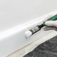 En hand med vit handske använder en pensel för att måla en vit golvlist.