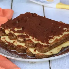 Tiramisù con crema pasticcera, strati di savoiardi e cacao in polvere su un piatto bianco, con un tovagliolo arancione sullo sfondo.
