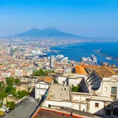A panoramic view of Naples, Italy, with Mount Vesuvius looming in the distance. The city's historic buildings and bustling port suggest that Naples is indeed nice.