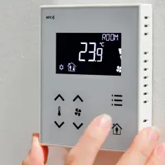 Dłoń dotyka panelu sterownika do powietrznej pompy ciepła, wyświetlającego 23.9°C.