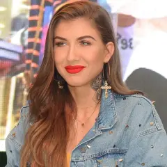 Deynn wiek, w jeansowej kurtce z ćwiekami, z wyrazistą czerwoną szminką i złotymi kolczykami w kształcie krzyża.