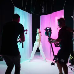 Filming a music video, learning how to color lighting with a dancer posed between blue and pink backdrops, while camera operators film.