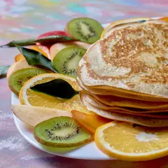 Stos naleśników z budyniem, otoczony plasterkami kiwi, pomarańczy i jabłka, ozdobiony listkami.