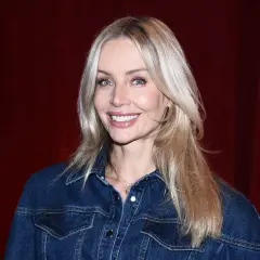 Uśmiechnięta blondynka z francuską grzywką w dżinsowej kurtce na tle czerwonej kurtyny.