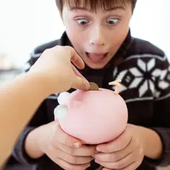 Un bambino con espressione sorpresa inserisce una moneta nel salvadanaio a forma di maialino, mentre una mano adulta lo aiuta. Sembra che il figlio stia rubando soldi ai genitori per il suo salvadanaio.