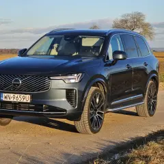 Ciemnoszary Volvo XC90 T8 na drodze polnej, z polami i drzewami w tle.