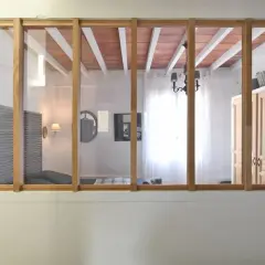 Une verrière en bois sépare deux pièces. On voit des volets, un lit, un lustre et des portes. Idéal pour savoir comment installer une verrière.