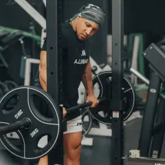 Mann mit Bandana macht schwere myo reps mit Langhantel im Fitnessstudio.