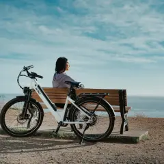 Kobieta odpoczywa na ławce obok białego roweru elektrycznego, podziwiając widok na ocean. Jaki rower elektryczny to świetny towarzysz podróży.