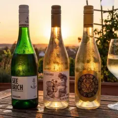 Drei Flaschen Wein und ein Glas stehen auf einem Holztisch. Im Hintergrund ein Weinberg bei Sonnenuntergang. Ein leichter Muskat Wein wäre hier perfekt.