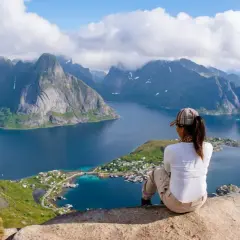 En person sitter på en klippa och blickar ut över en dramatisk kustlinje med berg och en by. En fantastisk vy för lofoten vandring.