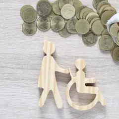 Drewniane figurki osoby pomagającej osobie na wózku inwalidzkim i monety. Czy zasiłek pielęgnacyjny wlicza się do dochodu?