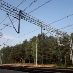 Trwa budowa sieci trakcyjnej nad torami kolejowymi, otoczona gęstym lasem sosnowym.