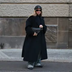 Kobieta w czarnym płaszczu oversize, czapce i okularach przeciwsłonecznych trzyma torebkę.