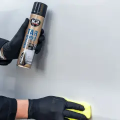 Ręce w czarnych rękawiczkach używają preparatu K2 Tar Remover, by usunąć klej z szyby auta.
