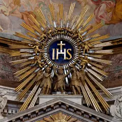 Goldene Strahlenkrone mit IHS-Symbol, das für Jesus steht. Putten und Engel umgeben das Symbol, das die göttliche Bedeutung von IHS unterstreicht.