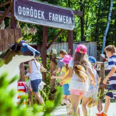 Dzieci biegną obok drewnianego szyldu "Ogródek Farmera" i figury krowy.