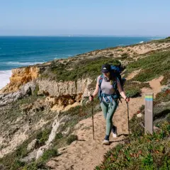 Wanderin auf dem Fisherman's Trail in Portugal. Eine Frau mit Rucksack und Stöcken wandert auf einem sandigen Küstenpfad mit Blick auf das Meer.