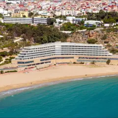 Widok na hotel przy piaszczystej plaży i turkusowym morzu, z miastem w tle.