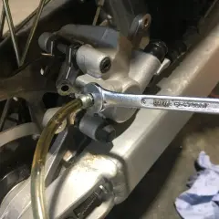 Klucz nasadowy odkręca odpowietrznik podczas wymiany płynu hamulcowego w motocyklu. Widać tarczę hamulcową i zacisk.