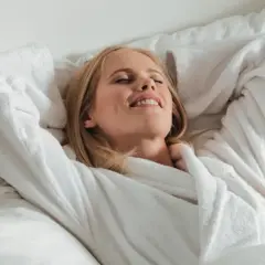 Frau entspannt im Bademantel im Bett, genießt den Moment. Vielleicht denkt sie über die richtige 5-HTP Dosierung nach, um sich noch besser zu fühlen.
