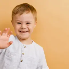Uśmiechnięty chłopiec z zespołem Downa macha ręką na tle w kolorze brzoskwiniowym.