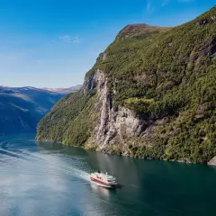 Ein Schiff fährt durch einen norwegischen Fjord im August. Das Wetter ist sonnig, die Berge sind grün und ein Wasserfall stürzt herab.