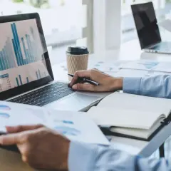 Mężczyzna w niebieskiej koszuli analizuje wykresy na laptopie, pracując jako **ekonomista**. Obok leży notatnik i kubek kawy.