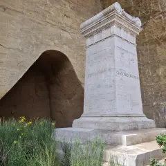 Monumento a Giacomo Leopardi nel Parco Virgiliano a Piedigrotta, con un'antica grotta sullo sfondo e fiori gialli.