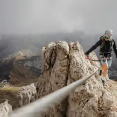 Kobieta na via ferrata polska pokonuje skalną grań, w tle górzysty krajobraz i kręta droga.