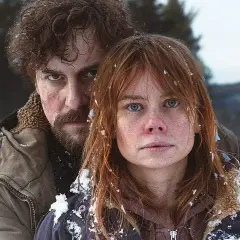 Julia Schlaepfer och en man med skägg och lockigt hår, båda med snö i håret, ser allvarligt in i kameran i en vinterlig skogsmiljö.