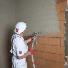 Pracownik w białym stroju nakłada tynk cementowo-wapienny na ścianę z pustaków. Czas schnięcia tynku zależy od warunków.