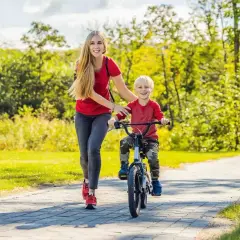 Mama pomaga synkowi, jak nauczyć dziecko jazdy na rowerze. Uśmiechnięci, na ścieżce w parku, wśród zieleni.