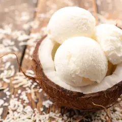 Tre palline di cremoso gelato al cocco servite in una noce di cocco su un tavolo di legno con scaglie di cocco sparse.