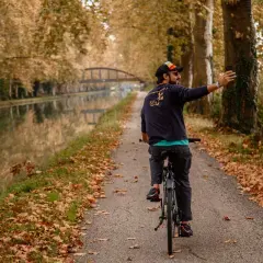 Hombre en bicicleta toma selfie en el Canal du Midi, rodeado de árboles otoñales y hojas caídas.