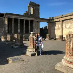 Pareja posa frente a ruinas antiguas, preguntándose donde está Pompeya. El sol ilumina columnas rotas y edificios históricos.