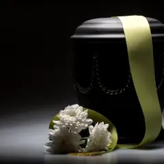 Czarna urna z zieloną wstęgą i białymi kwiatami. Rozważając koszty kremacji, warto pamiętać o tych symbolach.