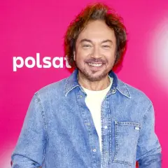 Mietek Szczęśniak, uśmiechnięty, w dżinsowej koszuli, na tle różowego banera z logo Polsat Hit Festiwal.