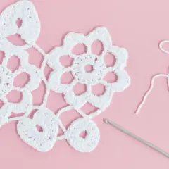 White Irish crochet lace with flowers and hearts, a crochet hook, and white yarn on a pink background.