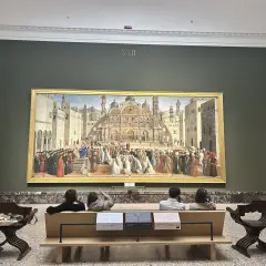 A grand painting depicting a historical scene is the centerpiece of this art in Milan gallery. Visitors sit on a bench, contemplating the detailed artwork.