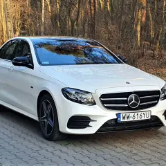Biały Mercedes E300e z czarnym dachem i lusterkami, zaparkowany na brukowanej ścieżce w lesie.