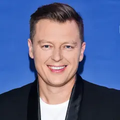 Rafał Brzozowski w czarnym garniturze na niebieskim tle z napisem "Bądźmy razem".