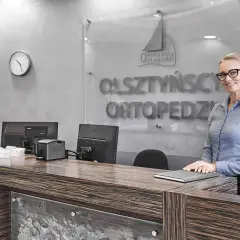 Recepcjonistka w okularach stoi przy ladzie w nowoczesnej klinice ortopedycznej.