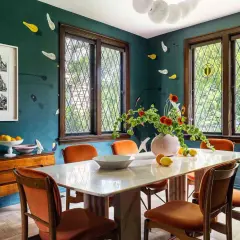 A vibrant kitchen and dining room design featuring a marble table, orange chairs, and a dark teal wall with playful accents.