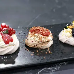 Tre små pavlovakakor med grädde och bär. En med hallon och vinbär, en med choklad och en med björnbär och pistage.