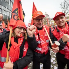 Drei Personen in roten Westen und Schals mit Gewerkschaftssymbolen, die Fahnen schwenken und die Faust ballen.