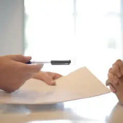 Dłonie trzymające dokument i długopis na tle jasnego okna.
