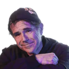 Andrea Satta, con un sorriso enigmatico, indossa un cappellino scuro e una felpa. La luce colorata crea un'atmosfera suggestiva.