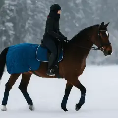 Kobieta w zimowym stroju odbywa jazdę konną zimą. Brązowy koń w niebieskim derce porusza się po ośnieżonym polu.