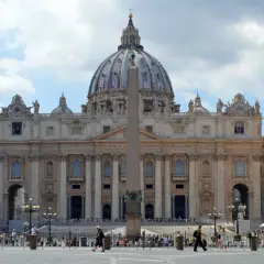 La maestosa facciata di San Pietro domina la piazza, con la sua cupola imponente e l'obelisco centrale. Turisti ammirano la grandezza.